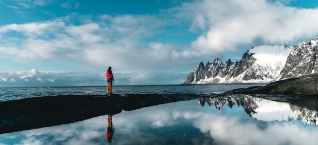 A person stands by a scenic snowy coastline reflected in still waters, under a vibrant blue sky.