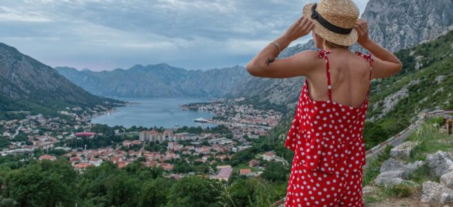 Back view of anonymous female enjoying picturesque scenery of rough rocky mountains and settlement near lake under cloudy sky